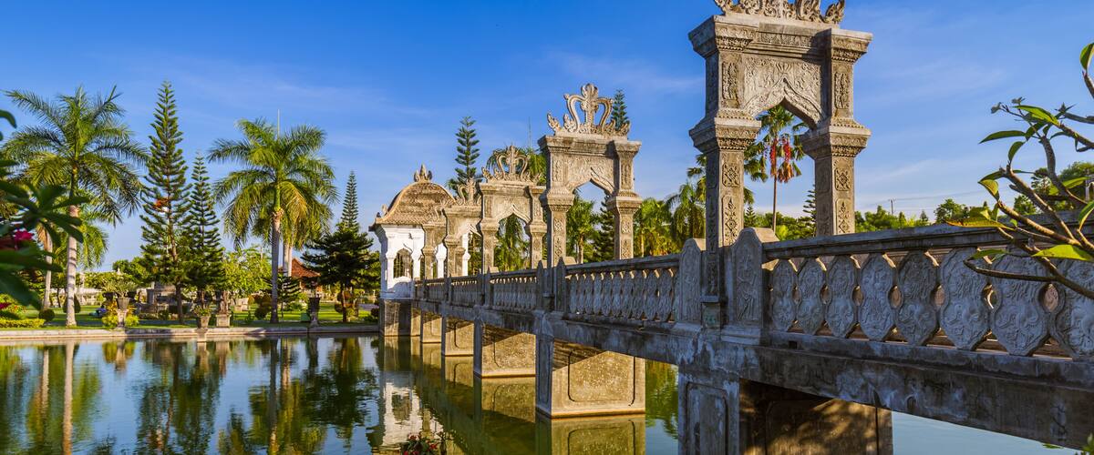 A stone bridge over a calm body of water with ornate pillars and palm trees in the background.