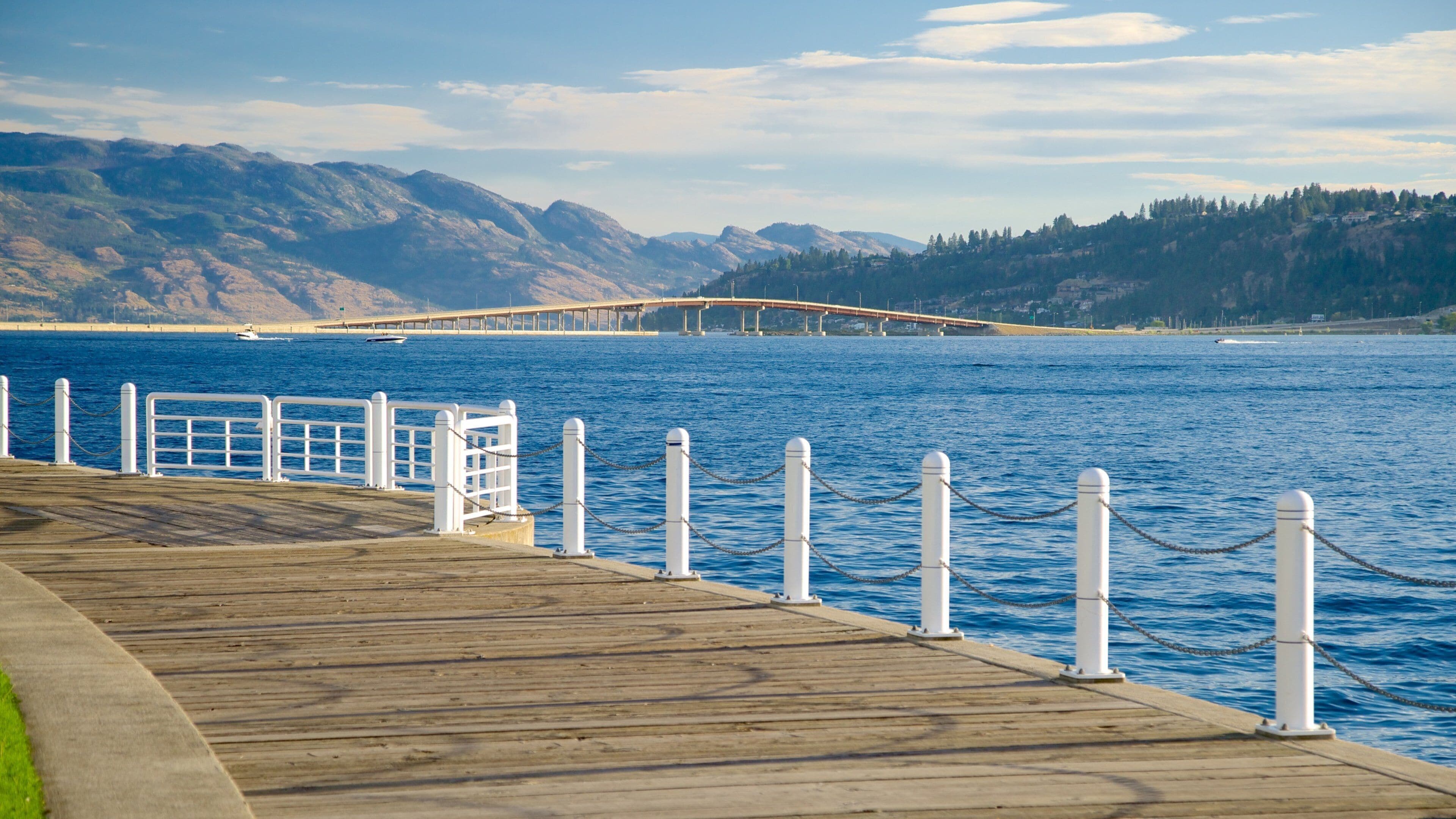 A wooden pier with white railings and bollards, leading to a body of water with a bridge in the background and mountains in the distance.