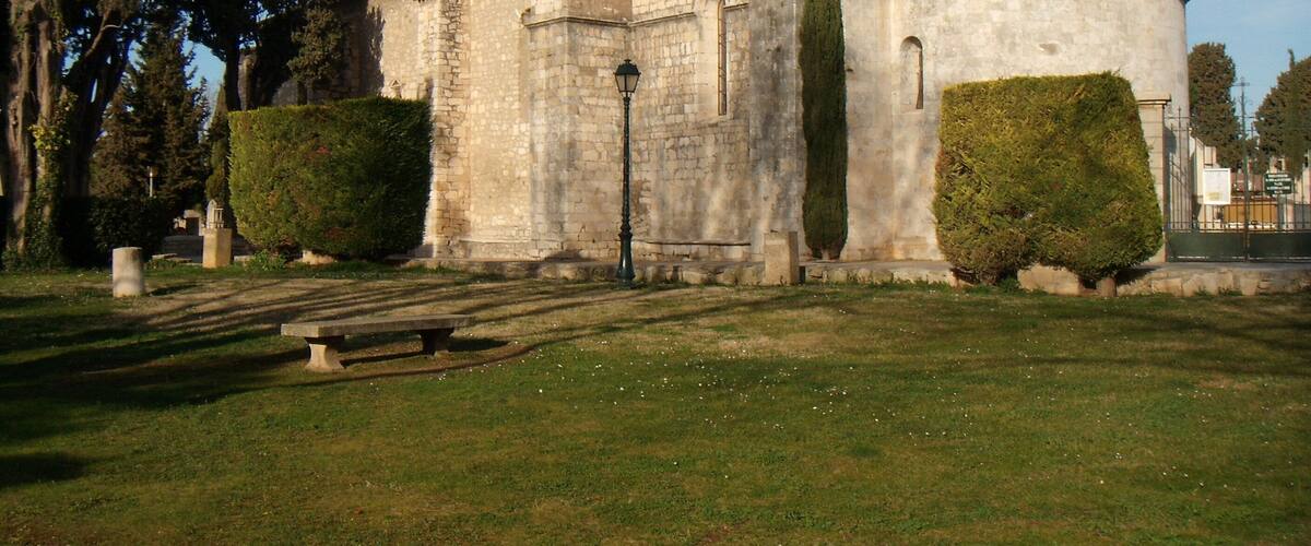 A historic stone church with a tiled roof, surrounded by well-maintained hedges and a grassy area with a bench.