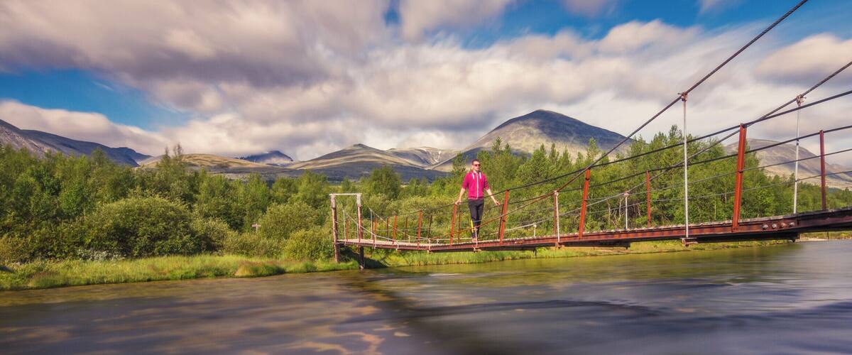A person walking on a suspension bridge over a river with mountains in the background.