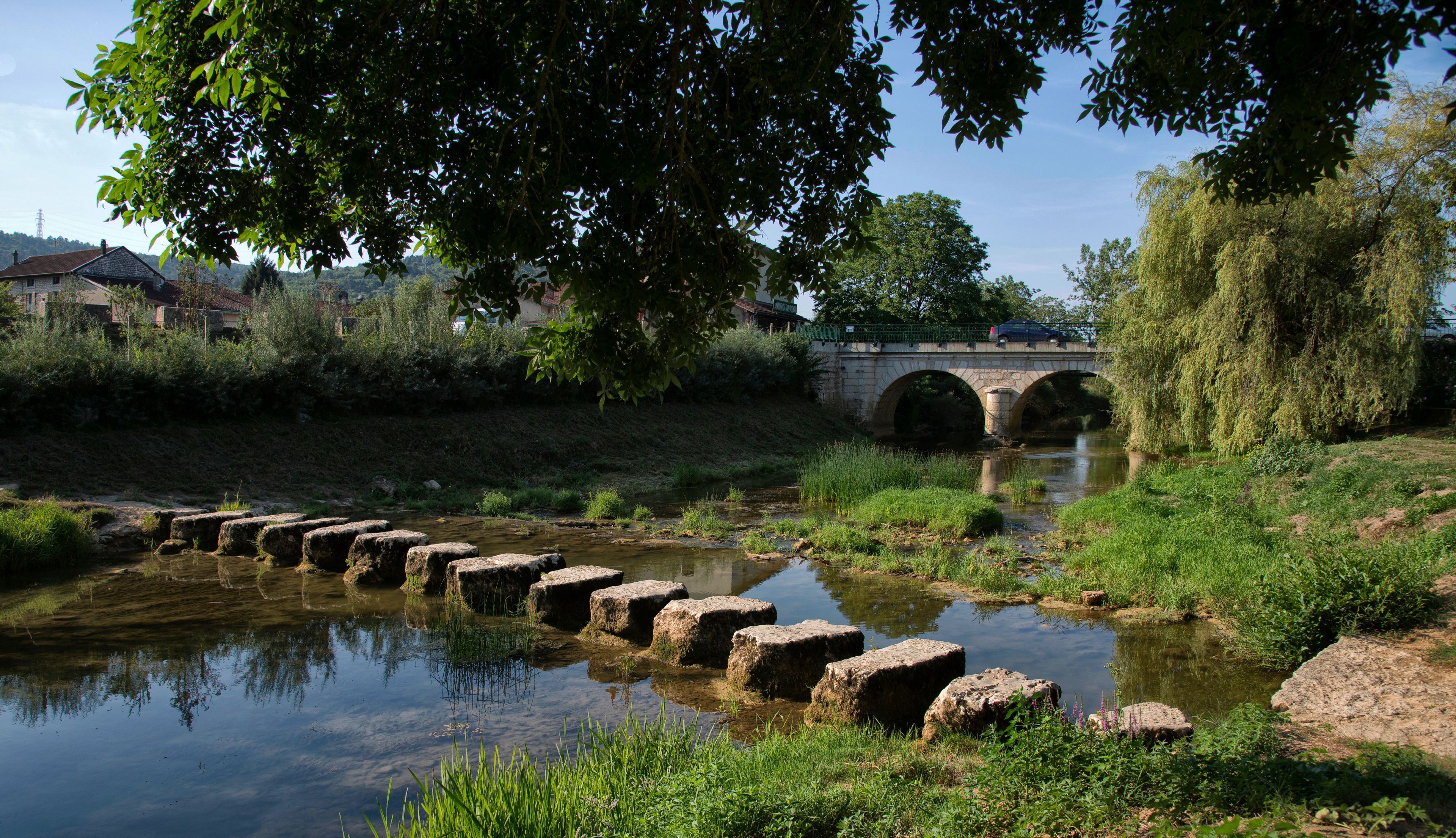 A stone bridge over a calm river, surrounded by lush greenery and a stone weir.