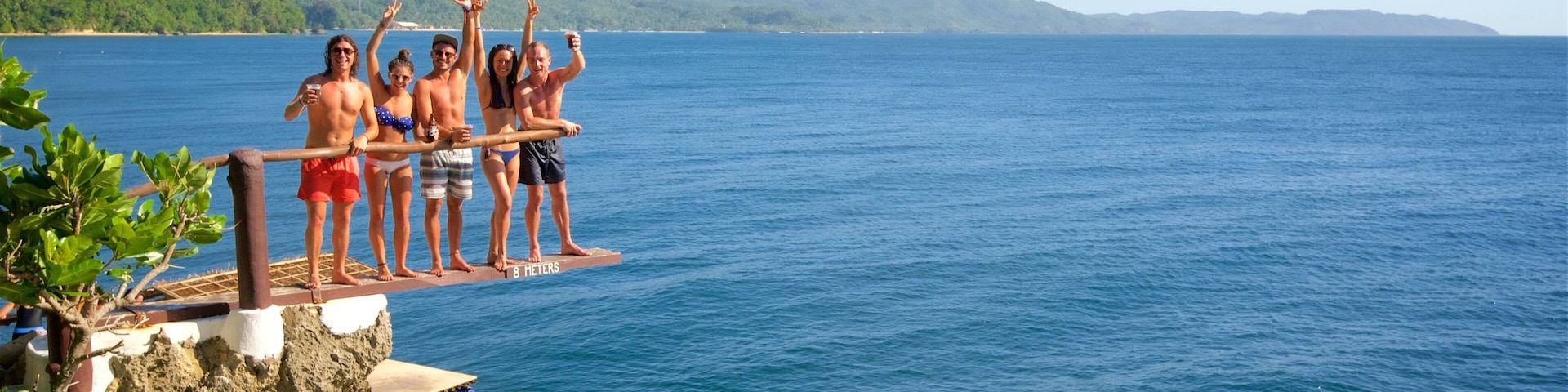 A group of people standing on a diving board over a body of water, with a boat visible in the background.