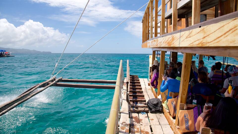 A wooden boat with passengers on a turquoise sea, a clear sky, and distant mountains.