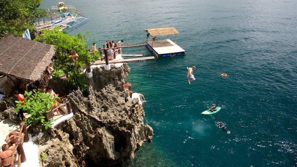A coastal scene with a rocky cliff, a dock with boats, and people engaging in water activities.
