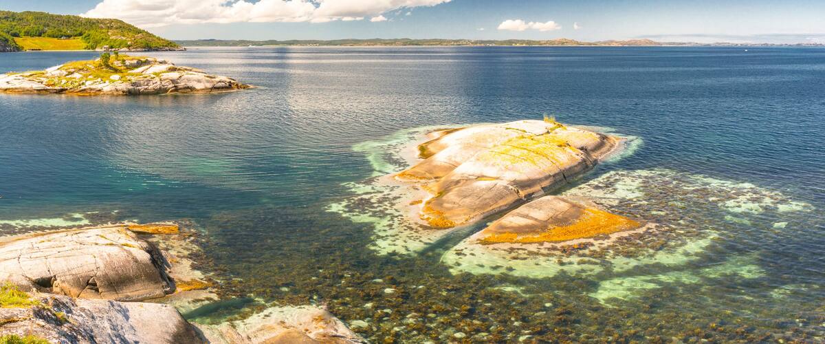 A coastal landscape with clear waters revealing underwater rocks and a small island with sparse vegetation.