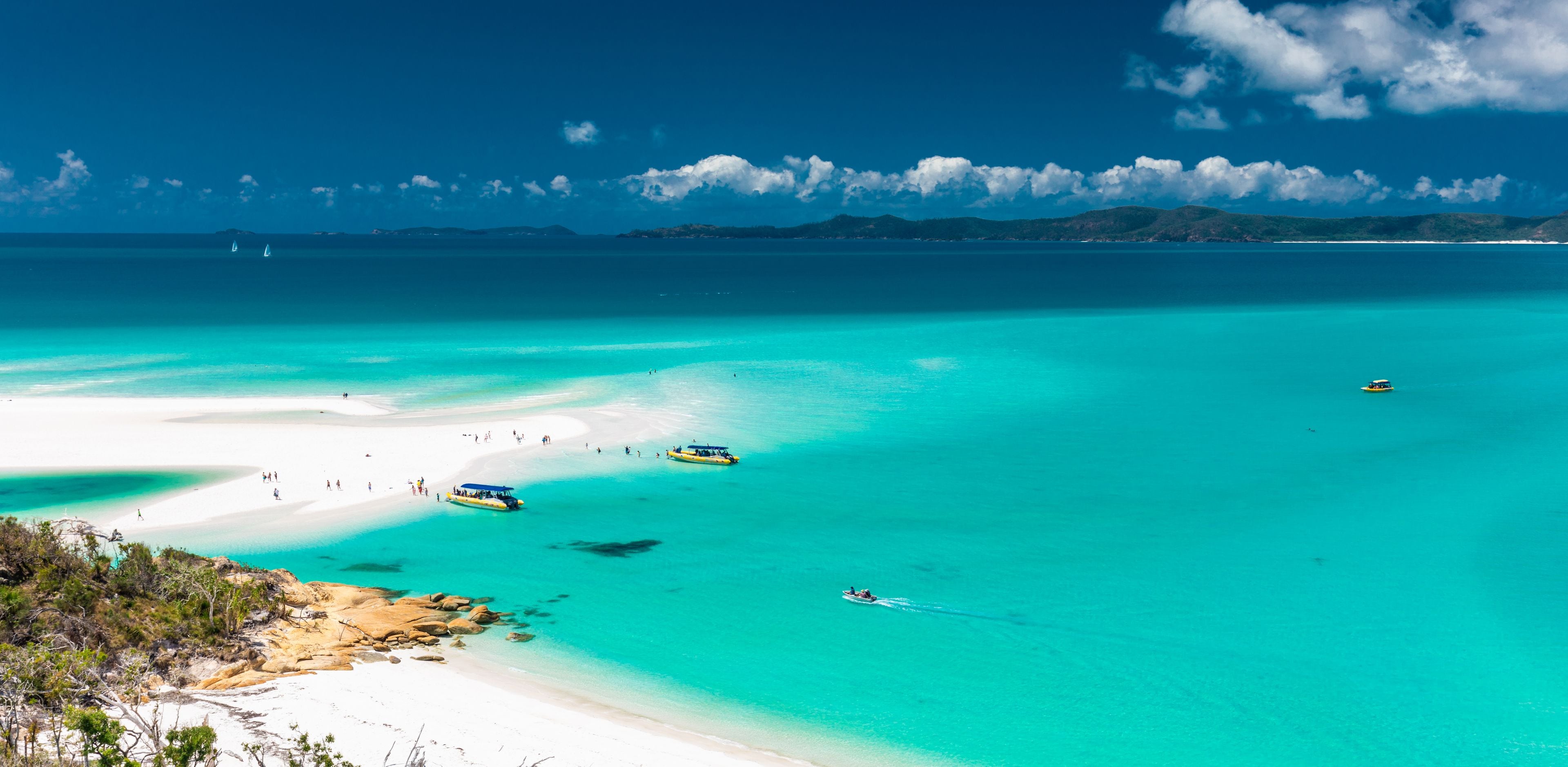 A turquoise sea with a white sandy beach, a few boats, and distant mountains under a clear sky.