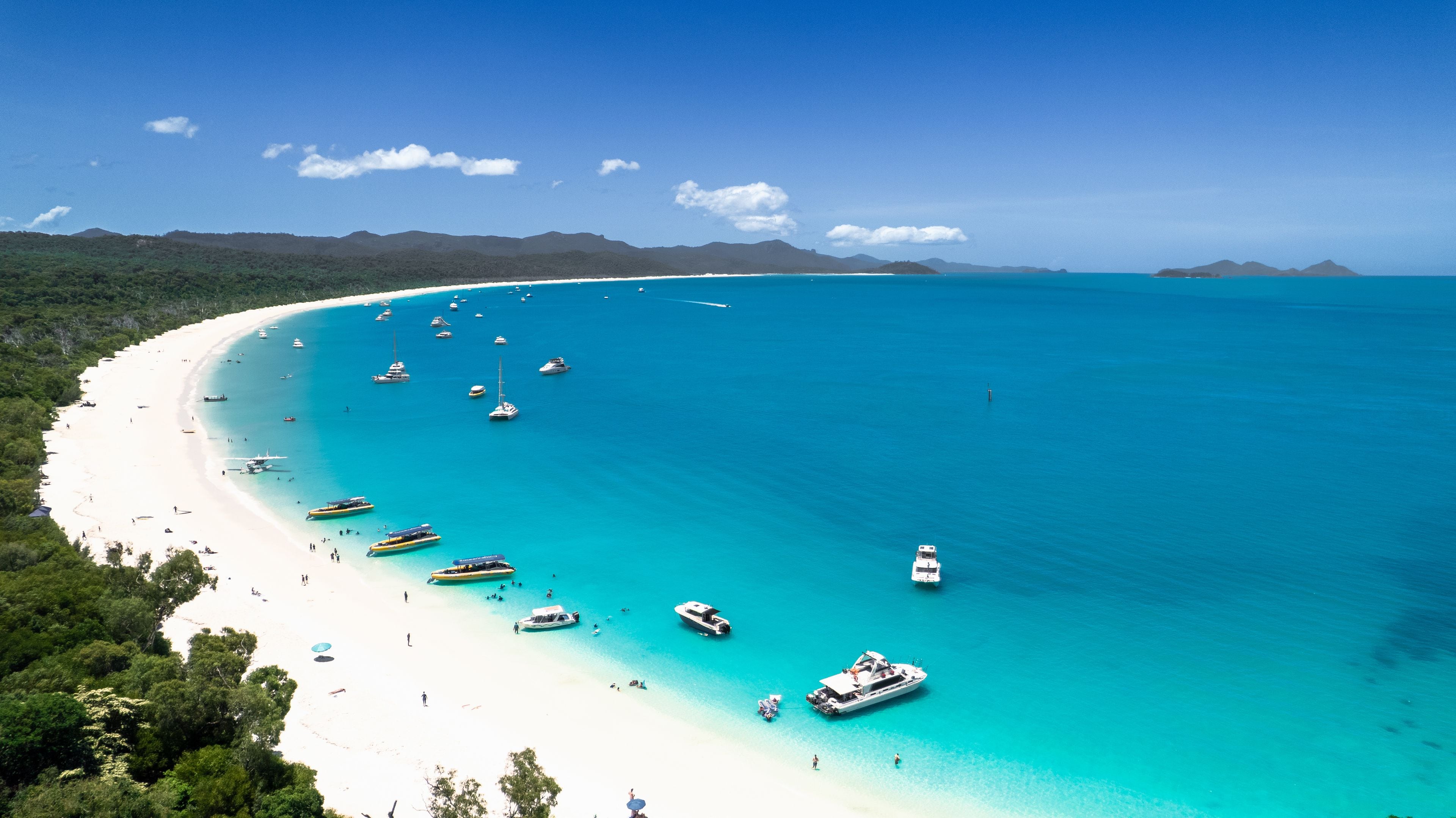 A coastal beach with clear turquoise waters, numerous boats, and a backdrop of green hills.