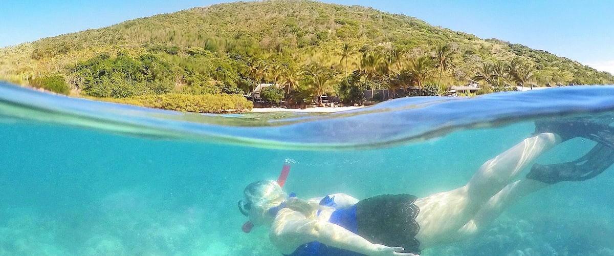 A person snorkeling underwater with a clear view of a coral reef and a hill with vegetation in the background.