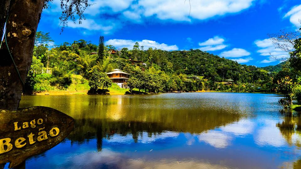A serene lake with a wooden sign reading "Lago Betão," surrounded by lush greenery and a clear blue sky.
