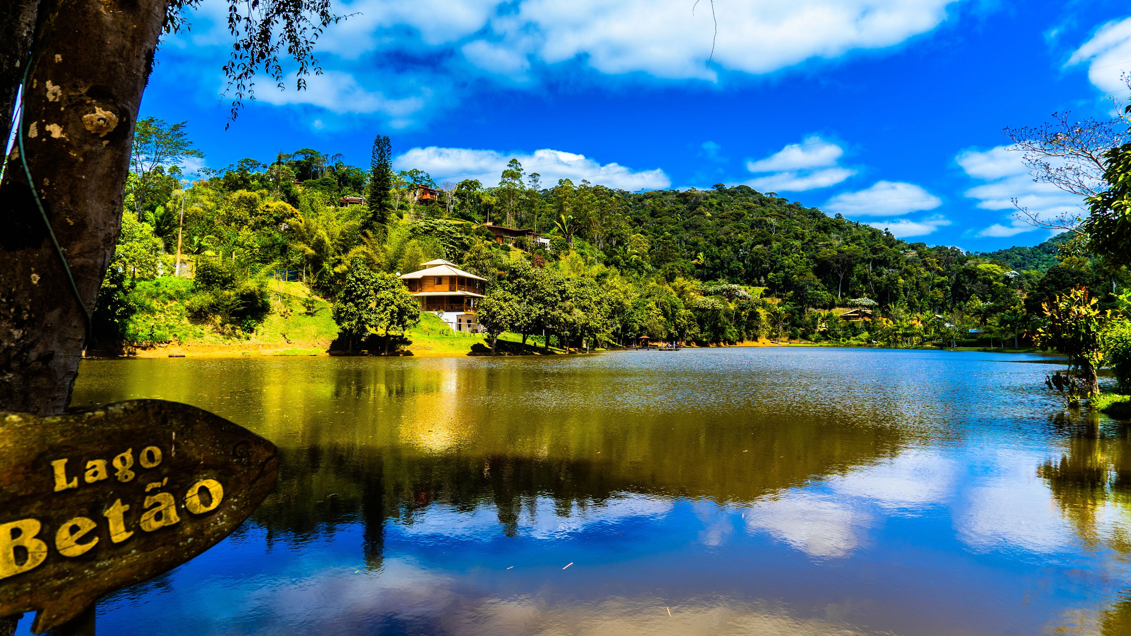 A serene lake with a wooden sign reading "Lago Betão," surrounded by lush greenery and a clear blue sky.