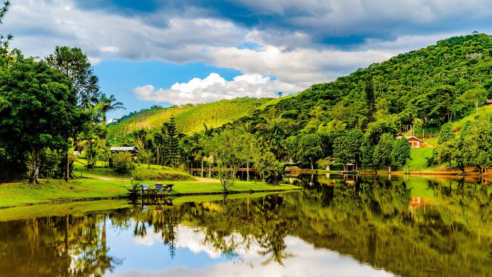 A serene lake surrounded by lush greenery and a hill with a small house.