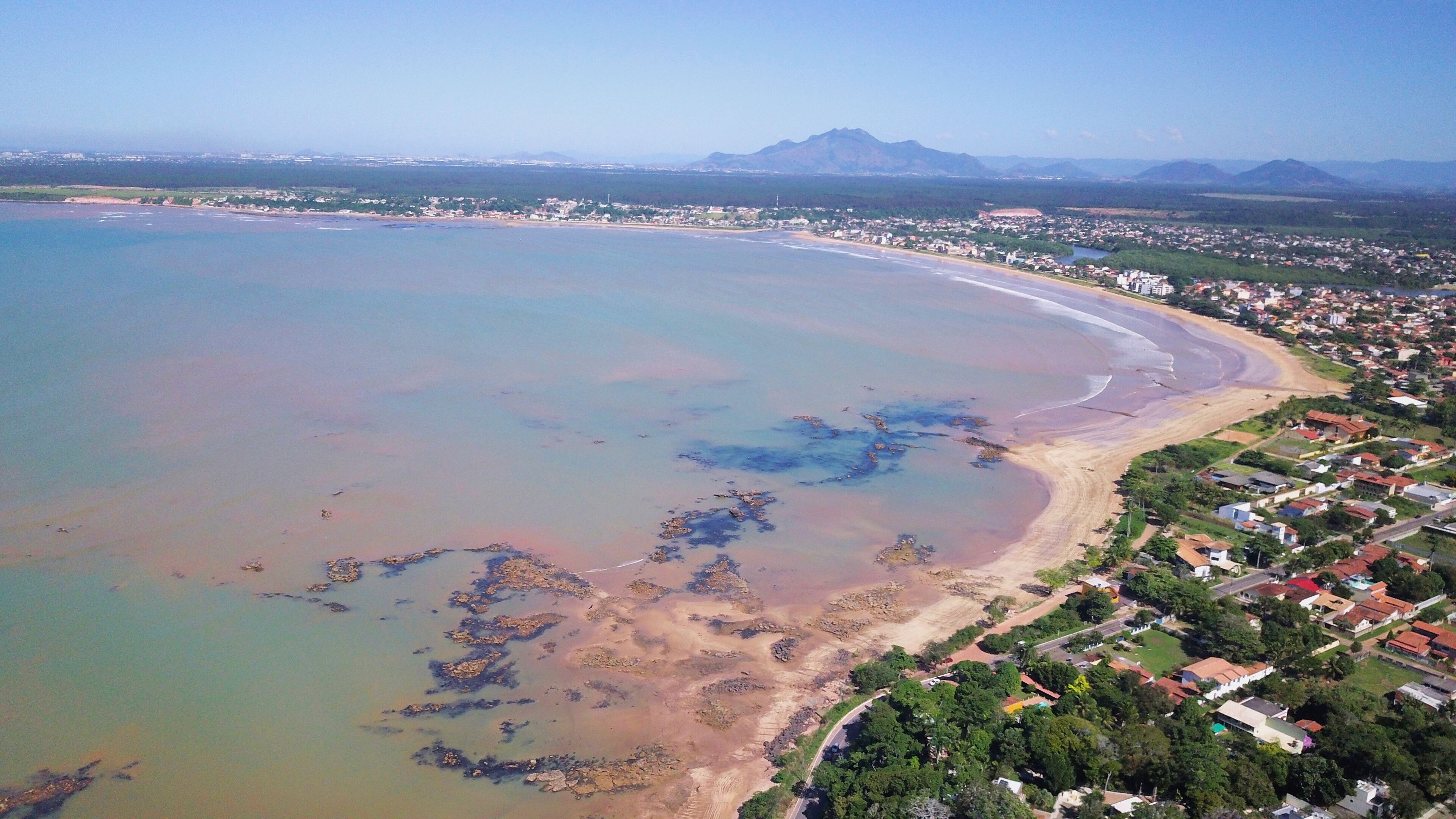 An aerial view of a coastal town with a beach, clear waters, and distant mountains.