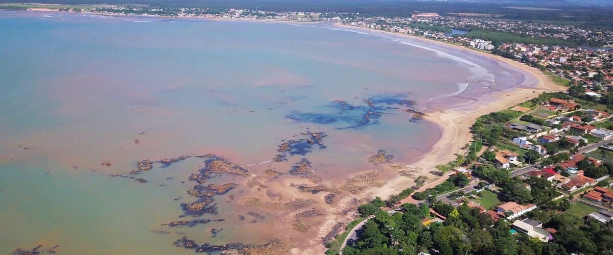 An aerial view of a coastal town with a beach, clear waters, and distant mountains.