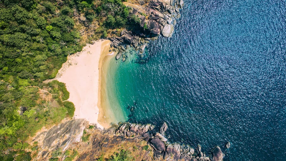 An aerial view of a coastal landscape with a sandy beach, clear turquoise waters, and surrounding rocky cliffs.