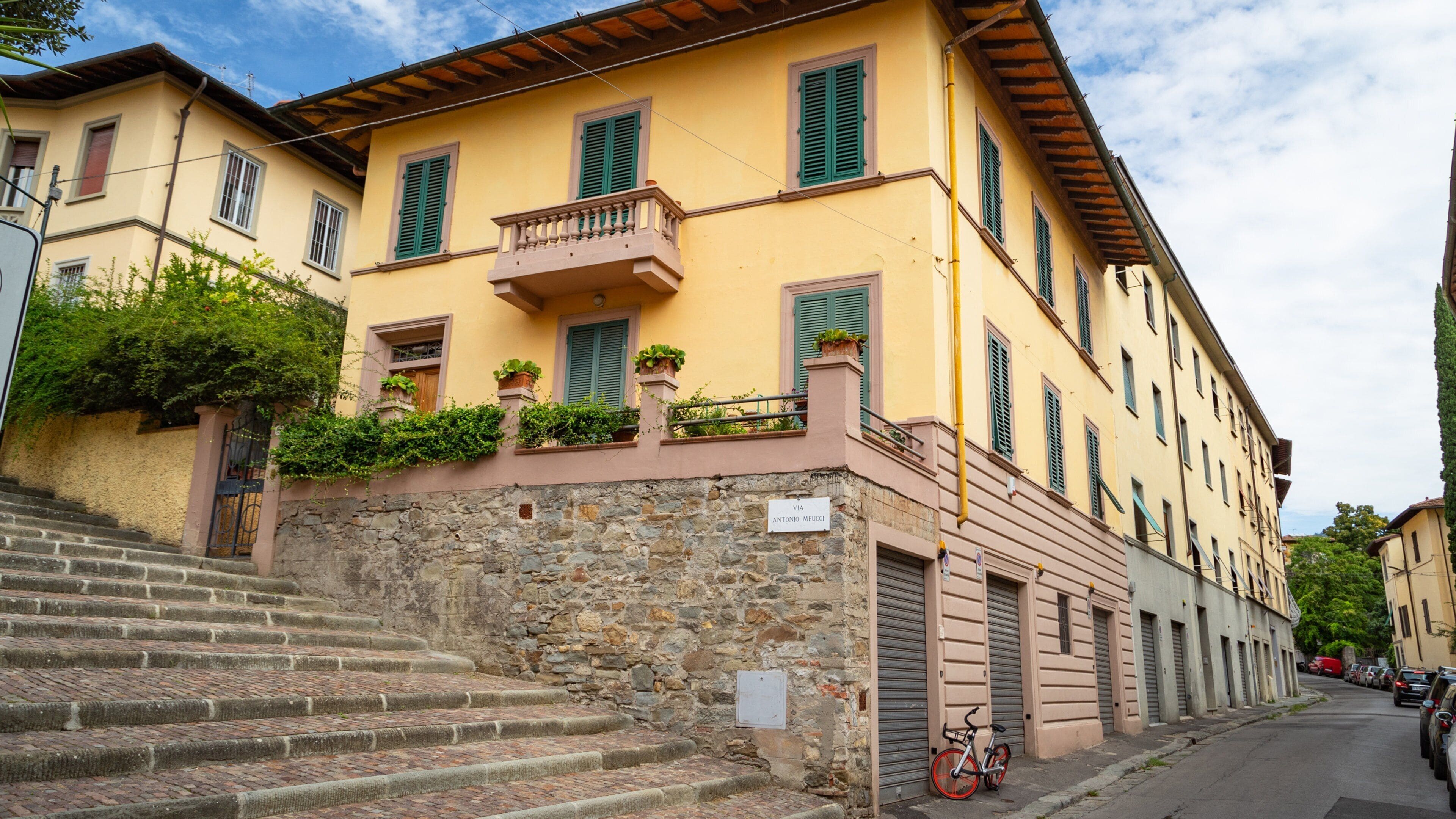 A yellow building with green shutters, stone steps, and a bicycle parked outside.
