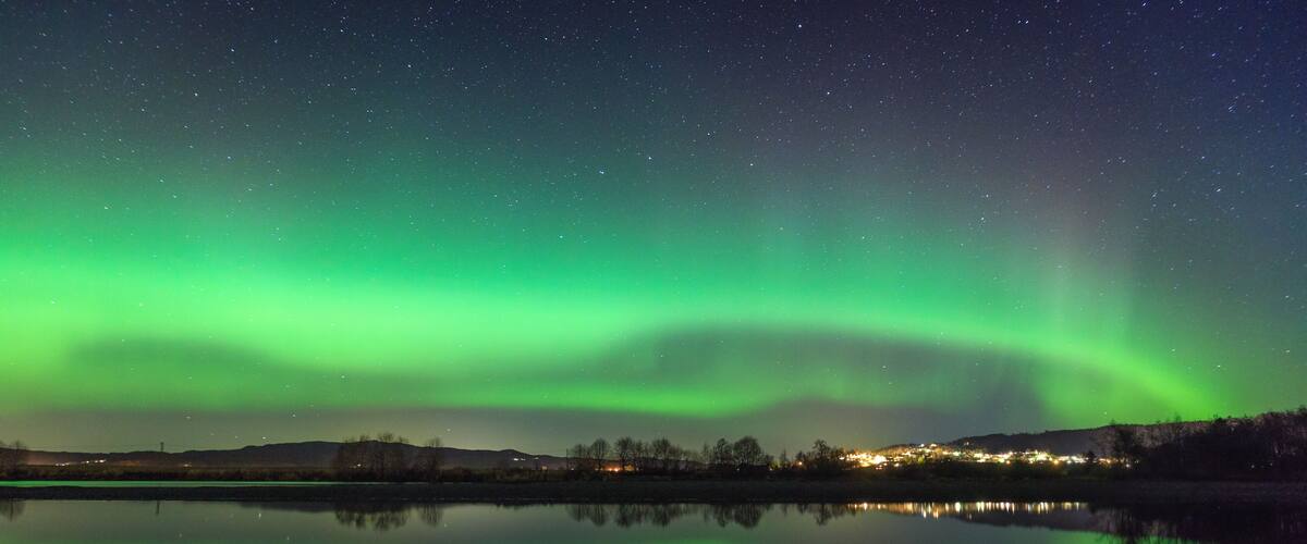 A night sky with the Northern Lights reflecting on a calm lake, with a distant town illuminated by lights.