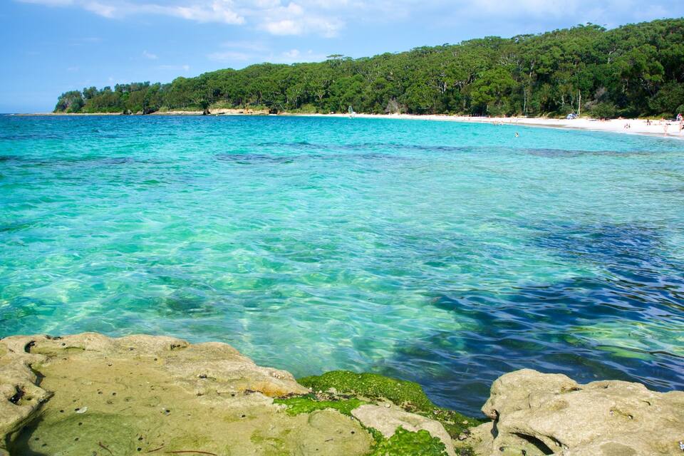 A clear turquoise sea with rocky foreground, a sandy beach, and a forested hill in the background.