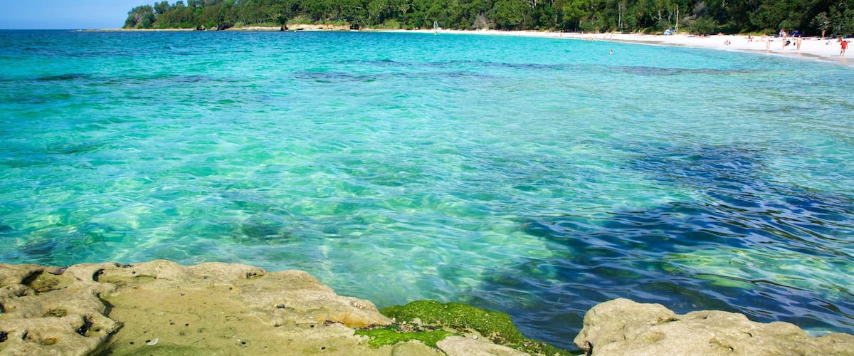 A clear turquoise sea with rocky foreground, a sandy beach, and a forested hill in the background.