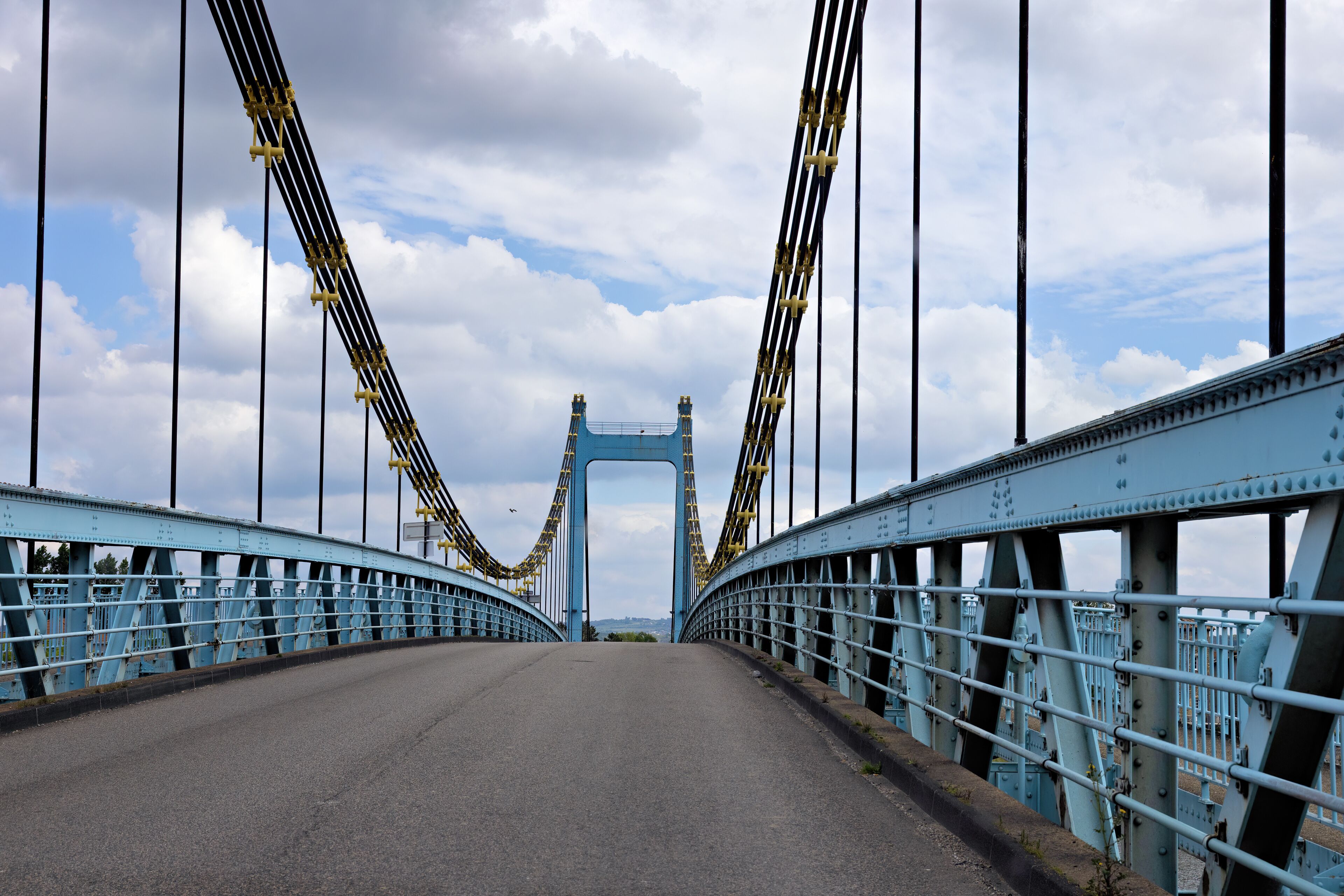 A blue suspension bridge with a clear sky and clouds in the background.