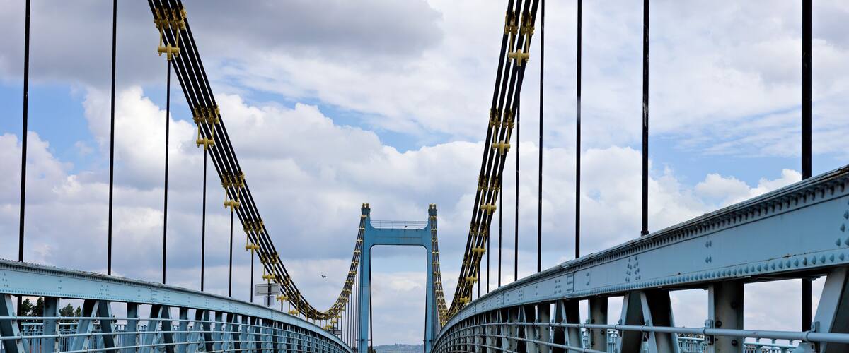 A blue suspension bridge with a clear sky and clouds in the background.