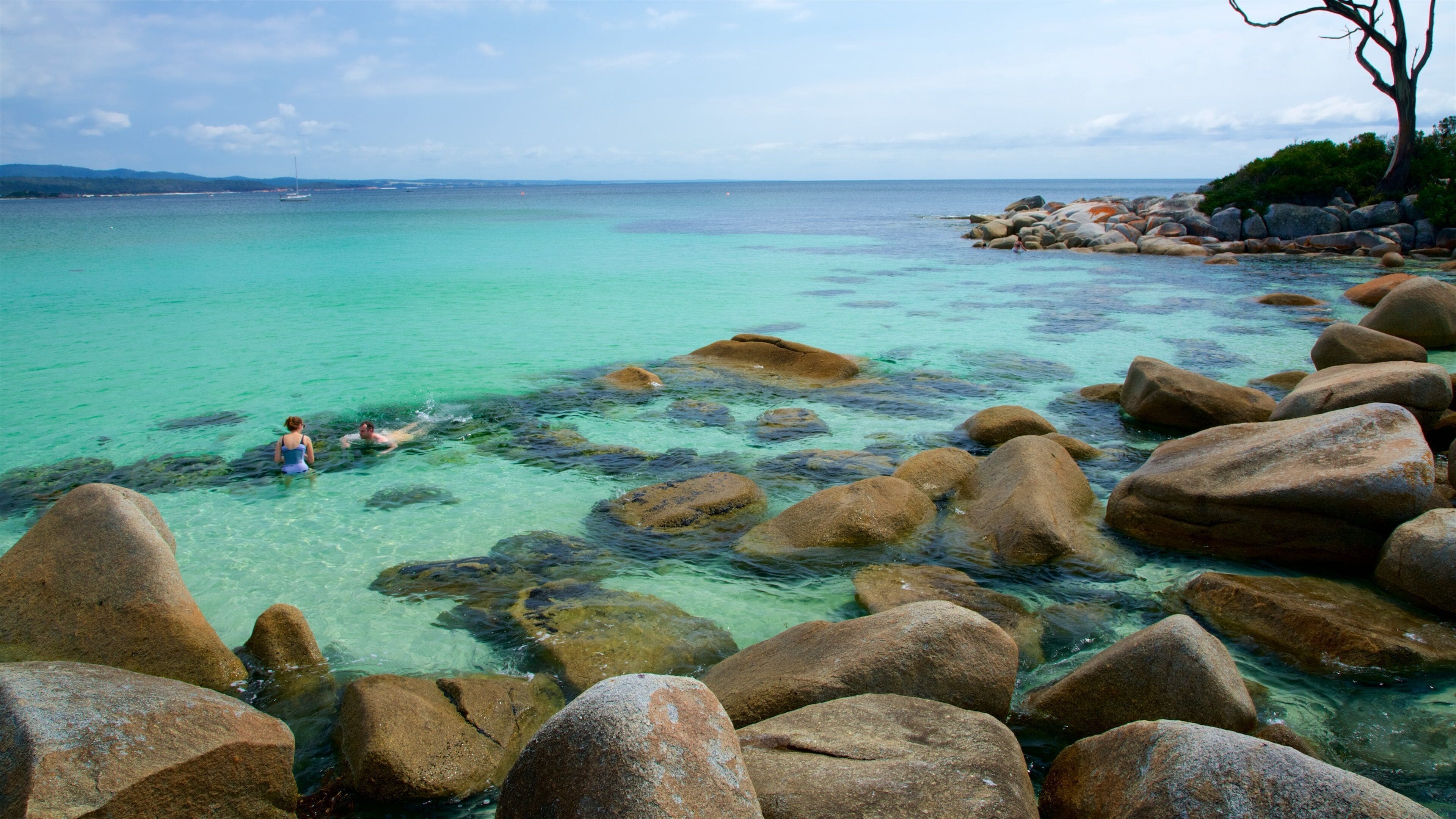 A clear turquoise sea with large rocks in the foreground and a person swimming in the water.