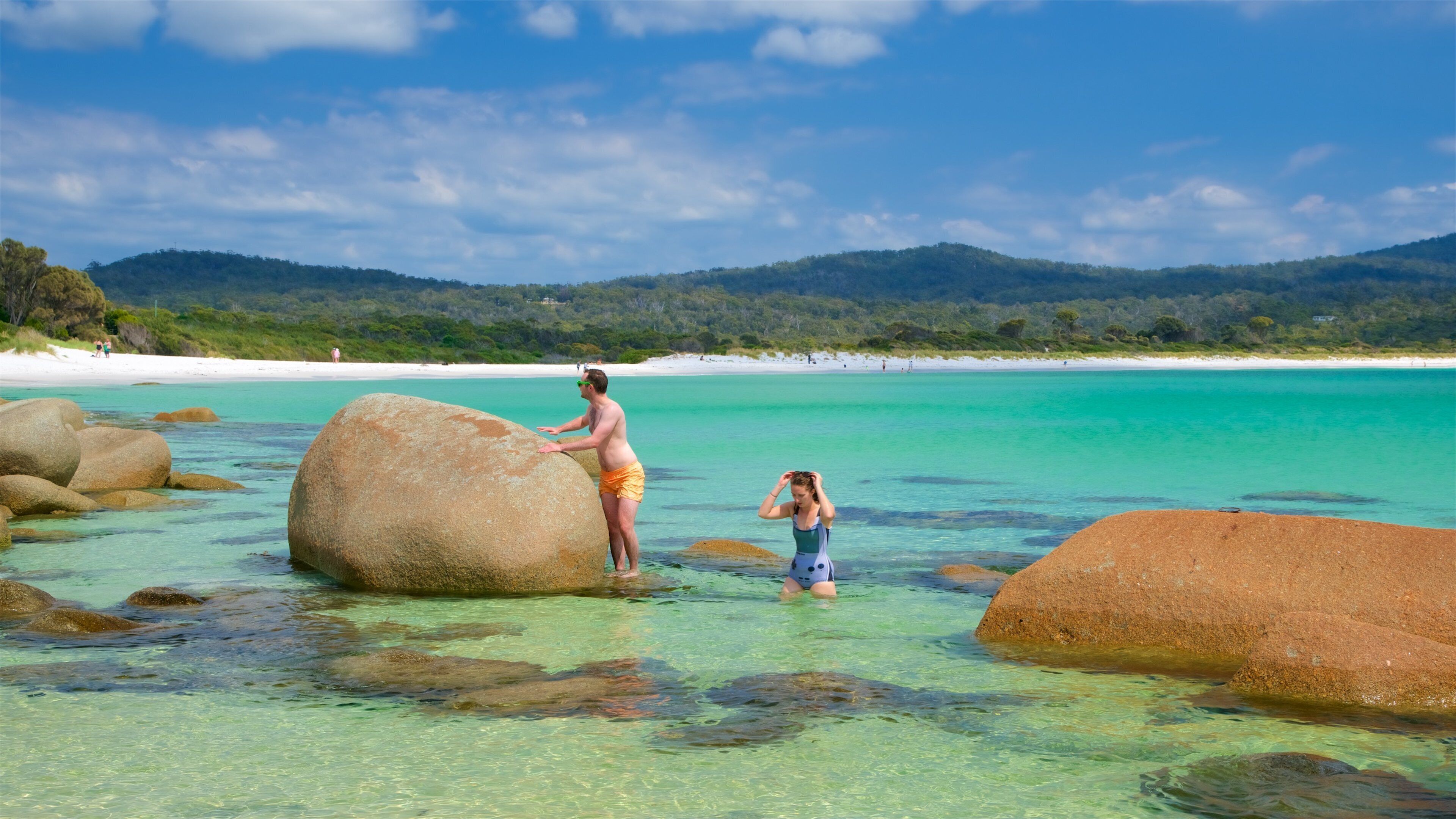 A beach with large boulders in shallow water, a clear blue sky, and distant hills.