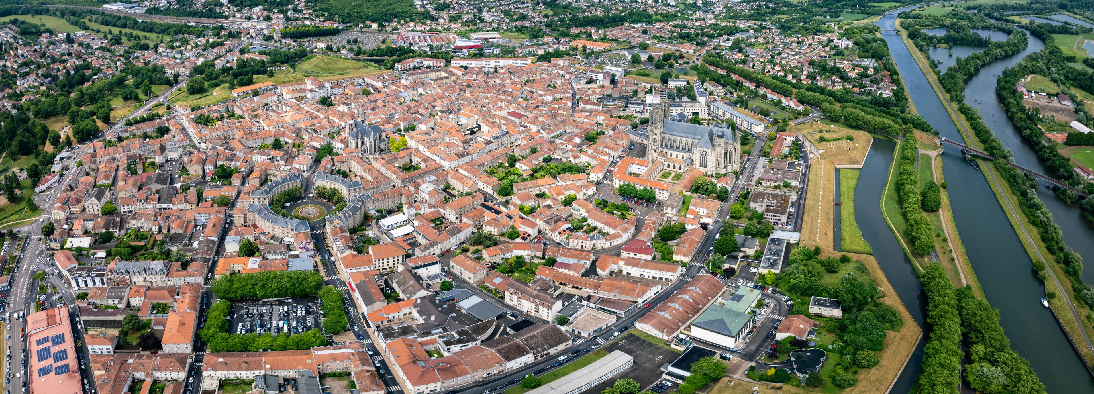 An aerial view of a historic city with a central roundabout, a river, and surrounding greenery.
