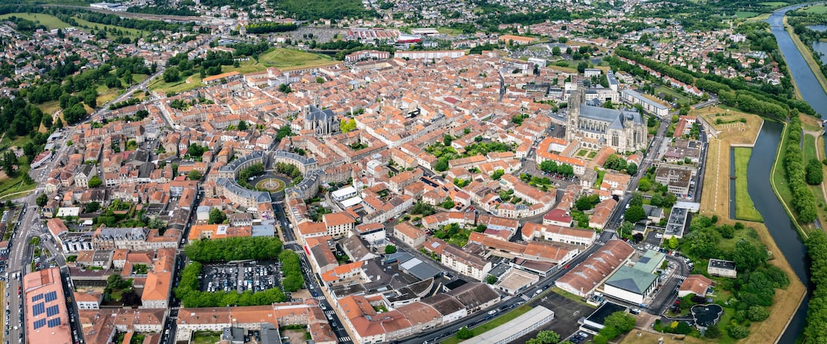 An aerial view of a historic city with a central roundabout, a river, and surrounding greenery.