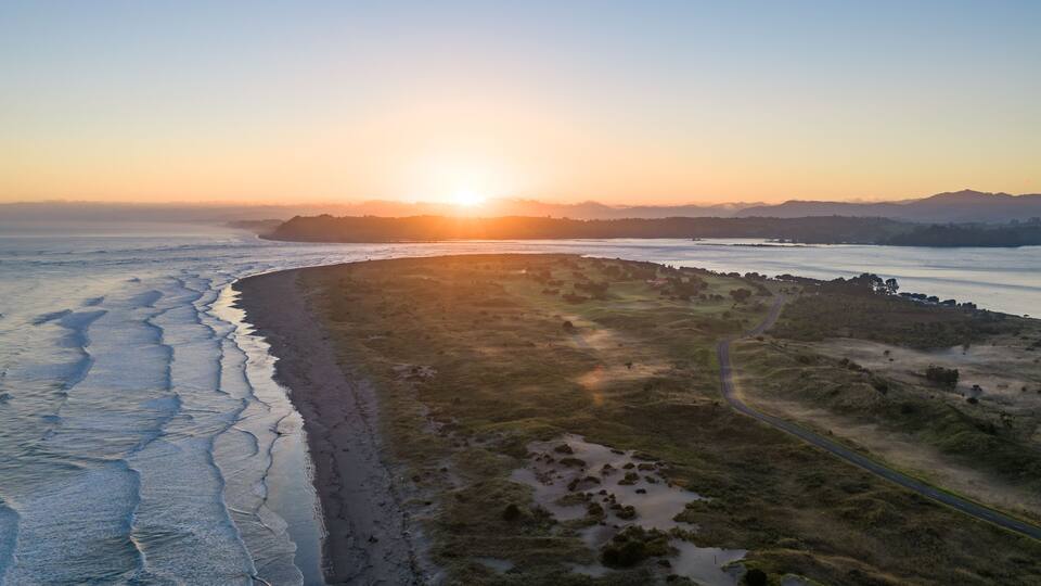 A coastal landscape with a winding road, a beach, and distant mountains during sunset.