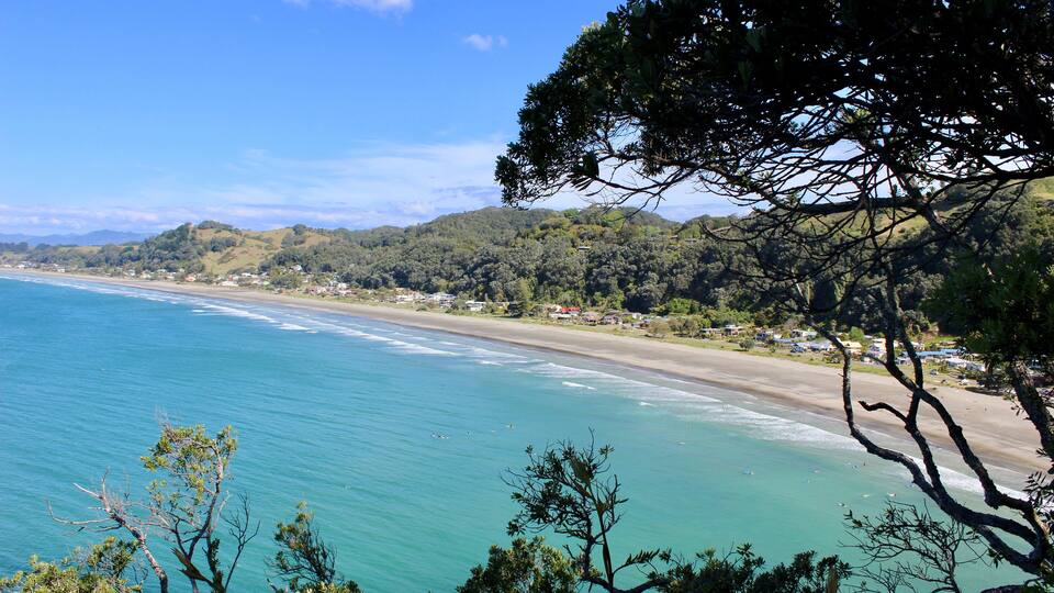 A coastal landscape with a beach, clear blue waters, and green hills in the background.