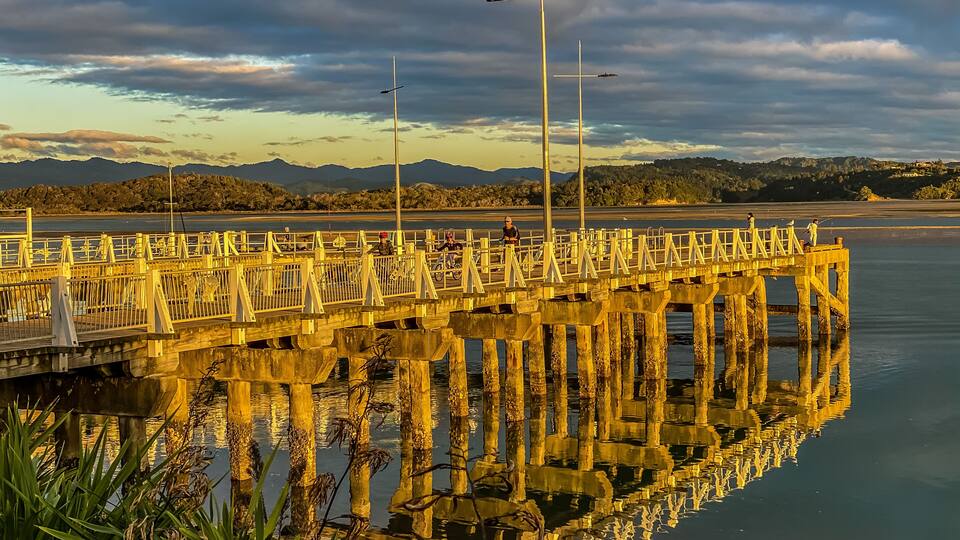 A wooden pier extending into calm waters, with mountains in the background and a cloudy sky.