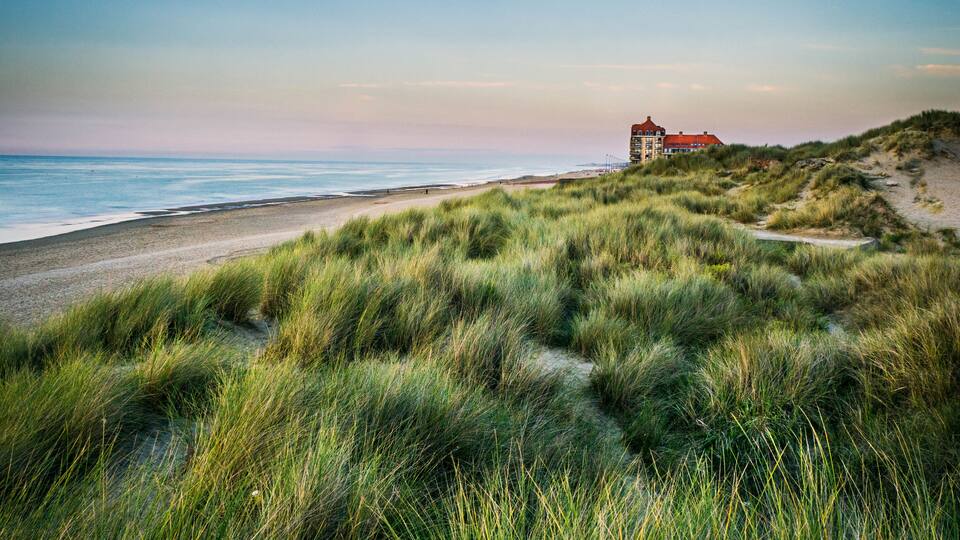 A coastal landscape with dunes, a beach, and a building on a hill.