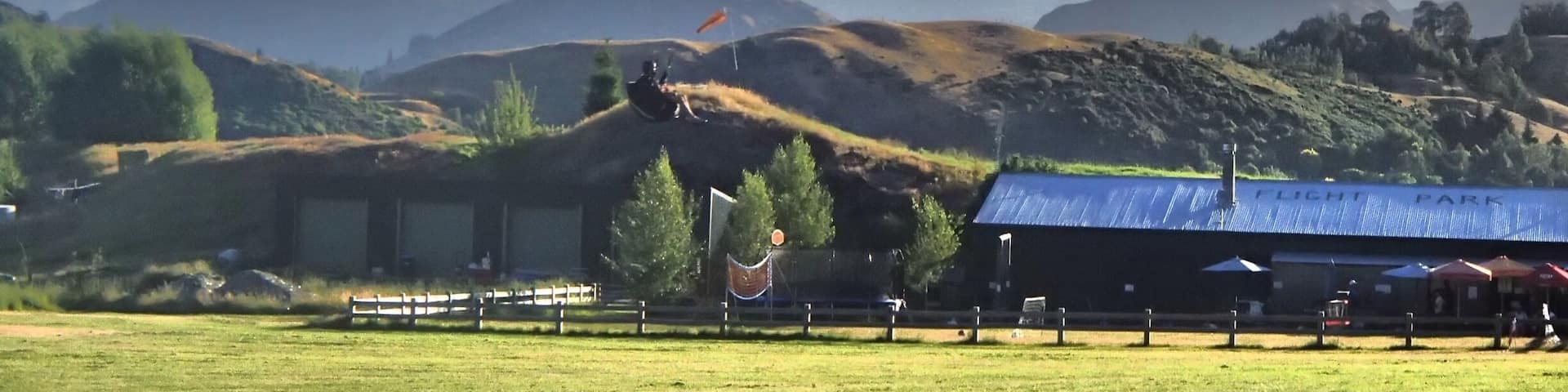 A person paragliding over a grassy field with mountains in the background and a building labeled "FLIGHT PARK".