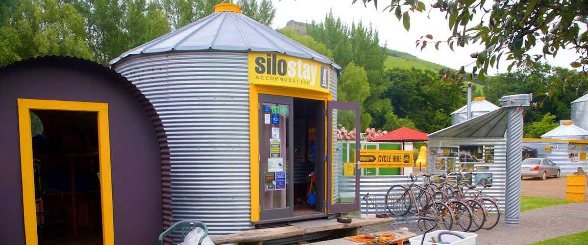 A small, yellow and purple accommodation building with a sign reading "Silostay Accommodation". There are bicycles parked outside and a small garden area with a wheelbarrow and a trash bin.