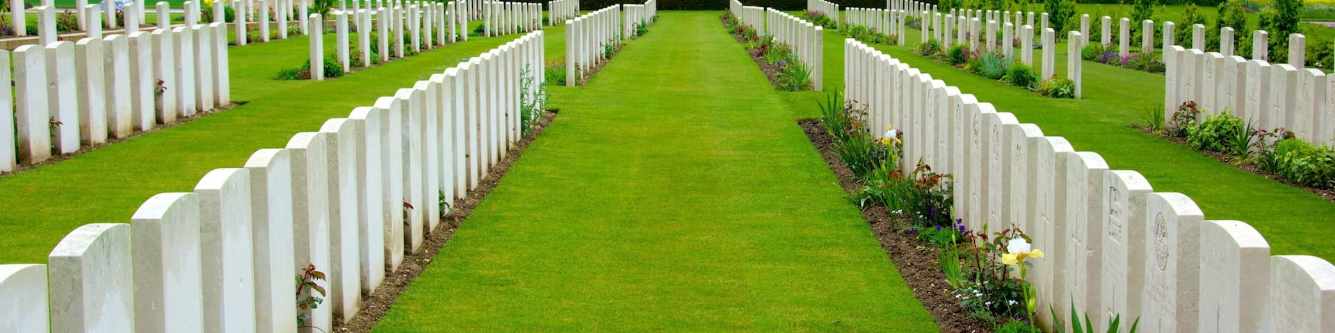 A cemetery with white headstones arranged in rows, a central building, and green fields in the background.