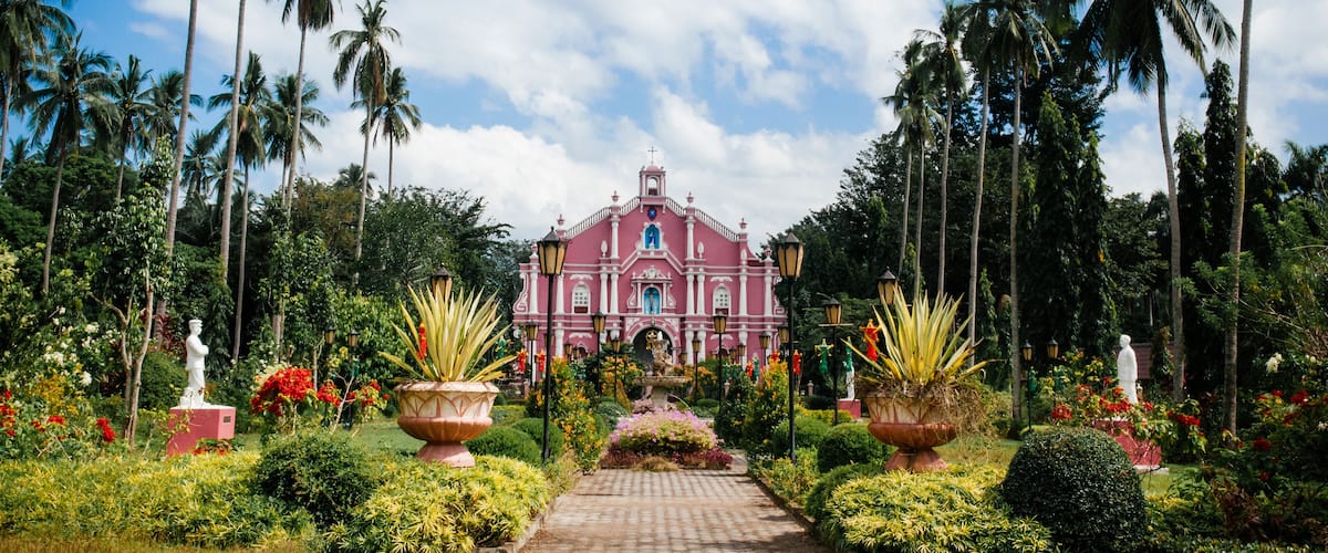 A pink colonial-style building surrounded by lush greenery and tall palm trees.
