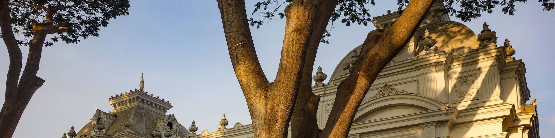 Un edificio clásico y majestuoso con columnas y estatuas ornamentadas, rodeado de un jardín cuidado con flores rosadas y un gran árbol.