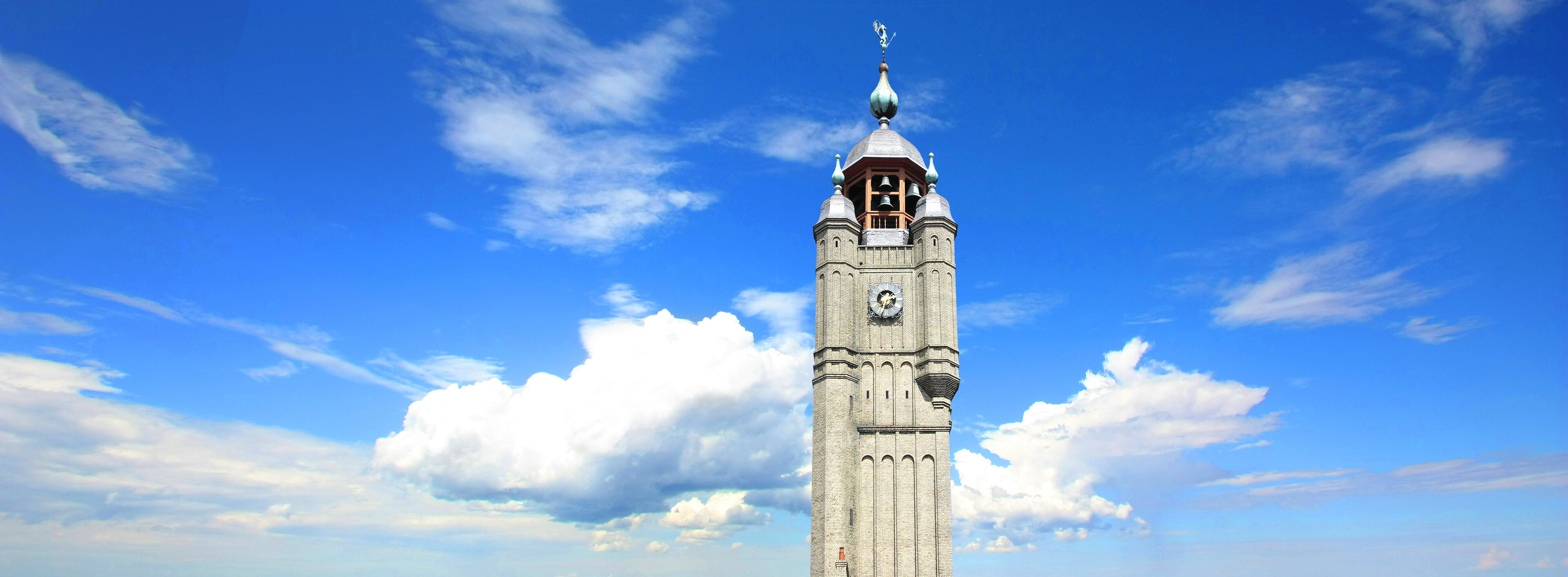 A tall clock tower with a pointed roof and a weather vane, set against a blue sky with scattered clouds.