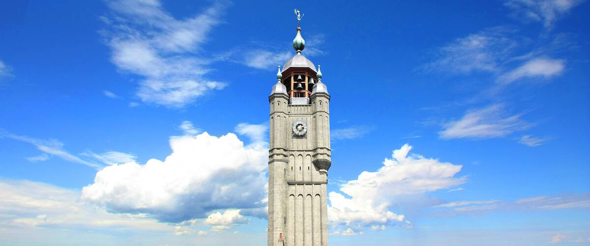 A tall clock tower with a pointed roof and a weather vane, set against a blue sky with scattered clouds.