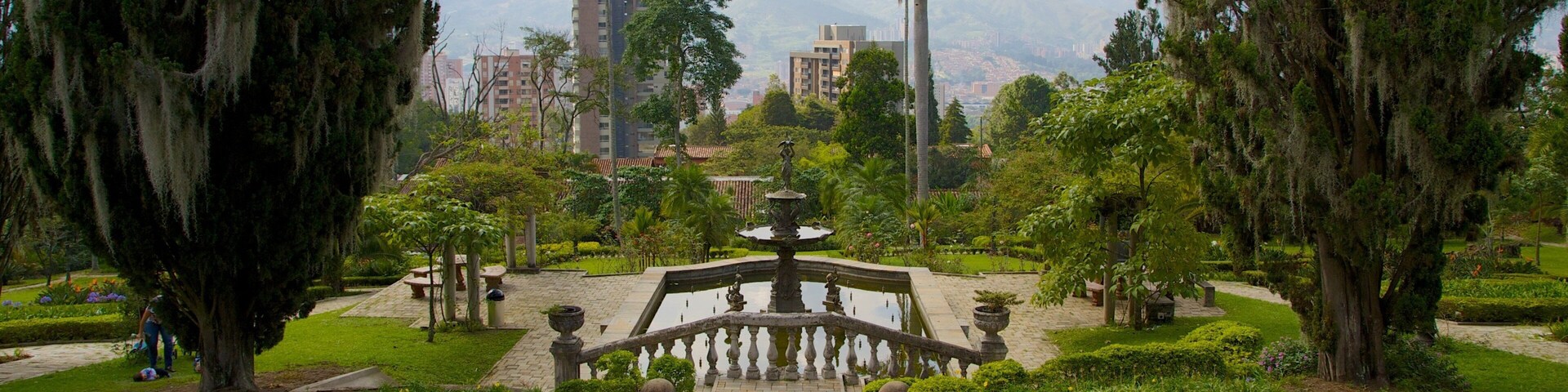 Um parque com uma fonte central, rodeado por relvados bem cuidados e árvores.
