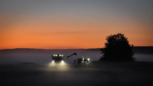 Combine harvester and tractor harvesting grains late at night in mist covered fields in the farmland of meteor impact crater Söderfjärden in Korsholm or Vaasa, in the region of Ostrobothnia, Finland.