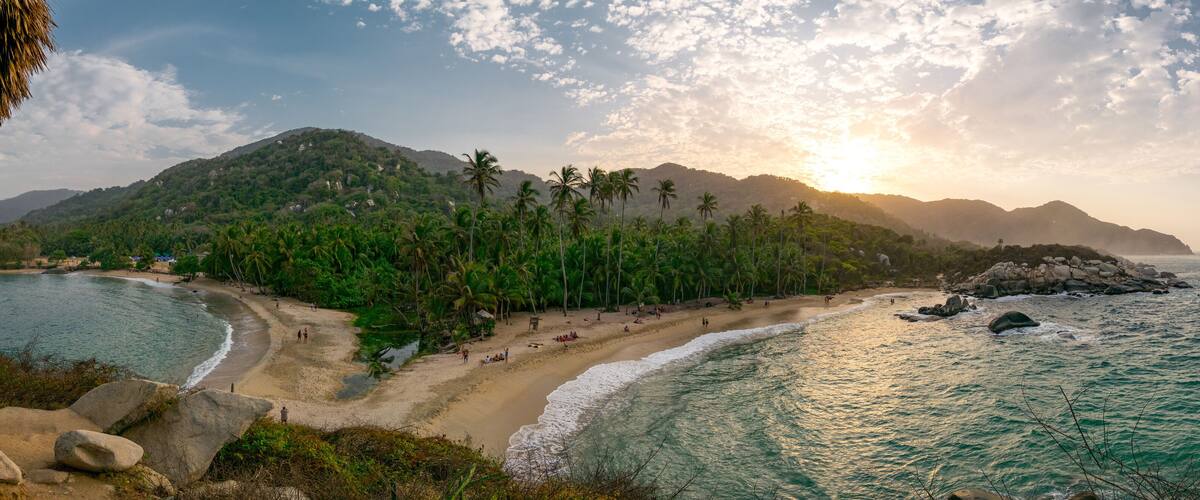 A beach with palm trees, a rocky outcrop, and mountains in the background.