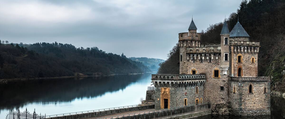 A medieval castle by a calm river, surrounded by forested hills.