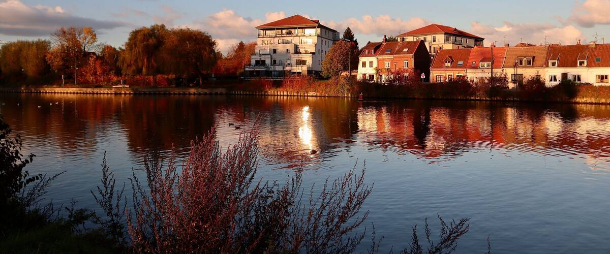 A calm river with buildings reflecting in the water, surrounded by autumn trees.