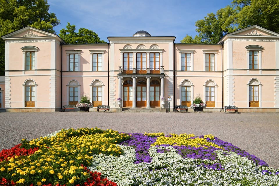 A large pink building with columns and arched windows, surrounded by a gravel driveway and colorful flower beds.