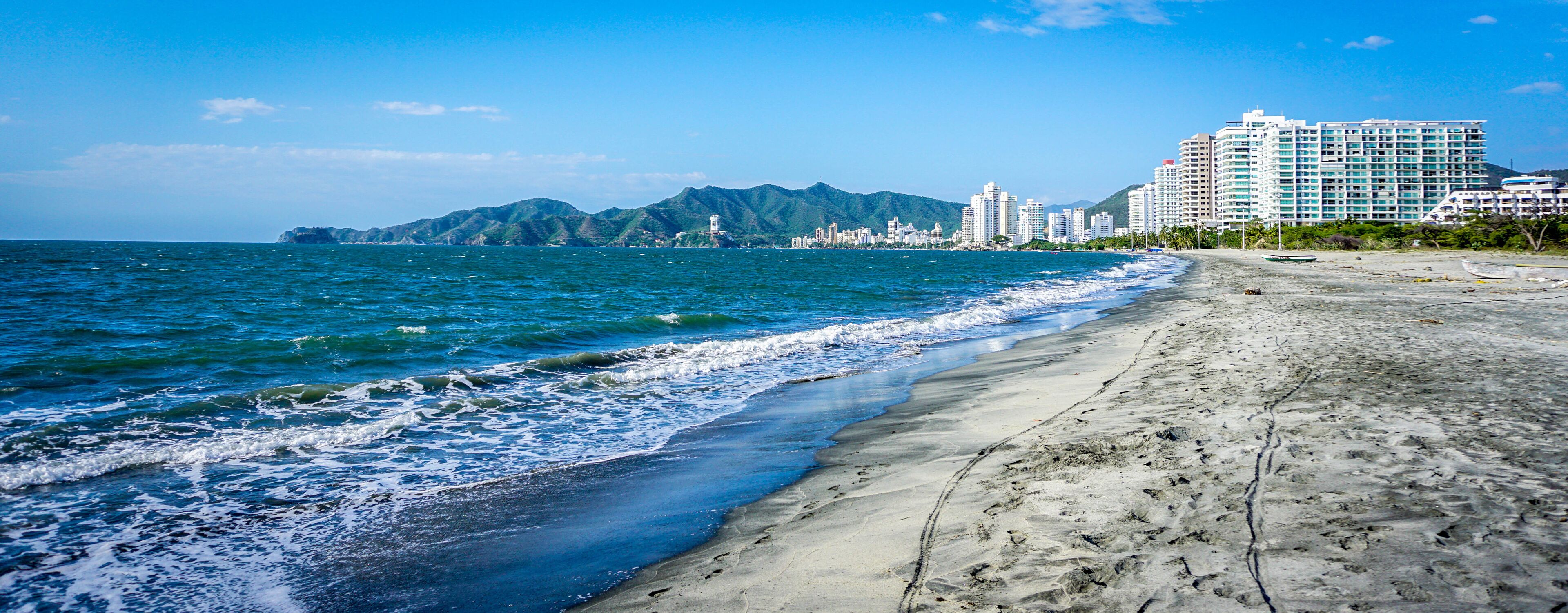 A beach with waves, a sandy shore, and a cityscape with buildings in the background.