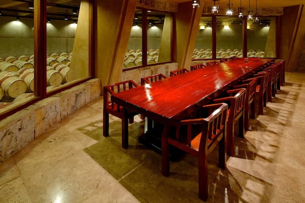 A long wooden table with chairs, a wine glass, and barrels in the background.