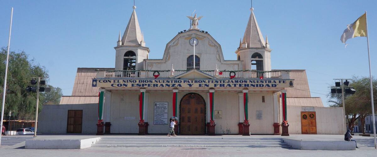 A church with two towers and a large central dome, featuring a sign that reads "Con el Niño Dios, Nuestro Patron, Festemos Nuestra Fe Con Esperanza y En Fraternidad".