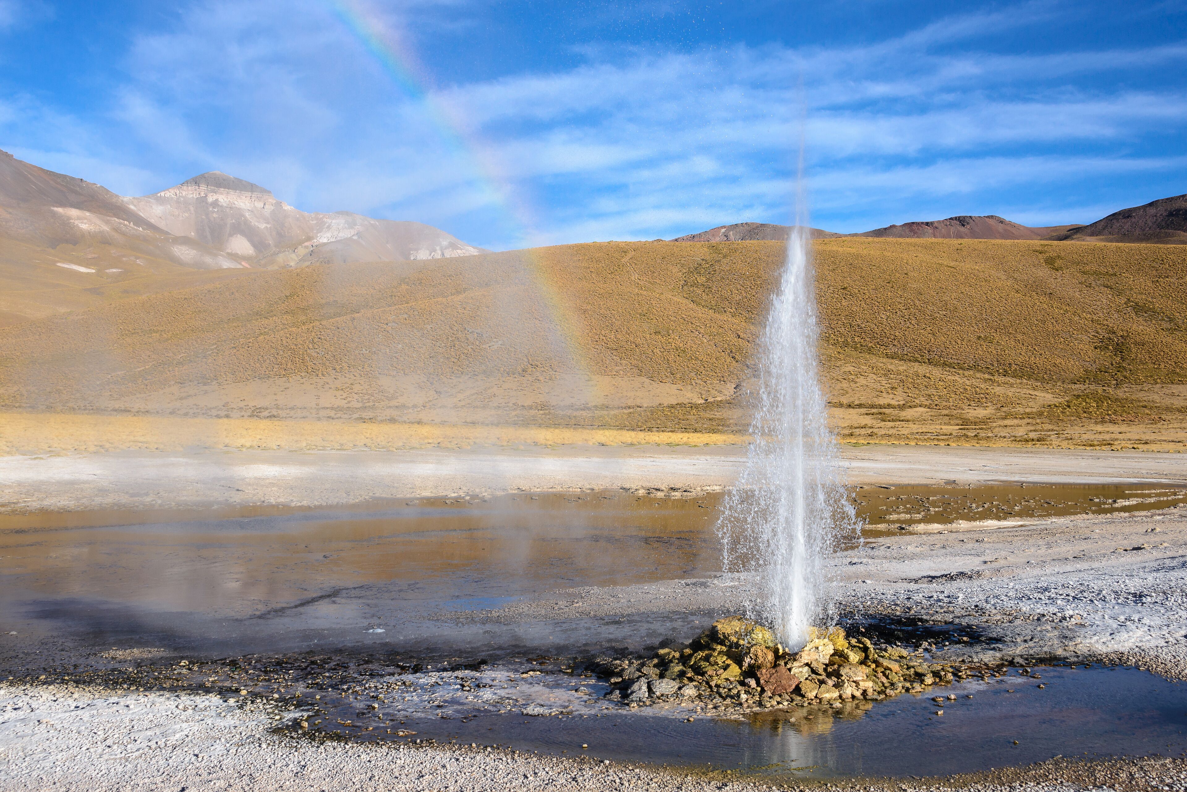 A geyser erupting in a dry, mountainous landscape with a rainbow in the sky.