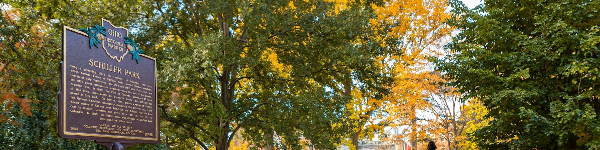 A park with a historical marker, trees with autumn leaves, and a paved path.